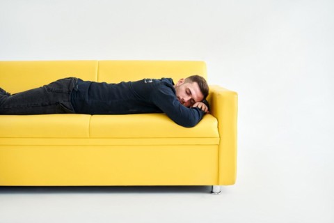 Tired young man lying down on yellow sofa over white background. Exhausted concept