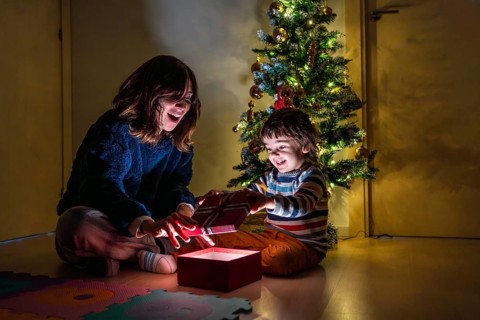 A young girl and boy are sitting on the floor in front of a Christmas tree. The girl is opening a