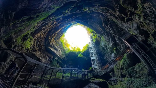 View of a large cave opening with stairs leading up, surrounded by lush greenery and rock