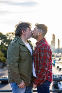 A gay couple, is standing on a city bridge while kissing each other