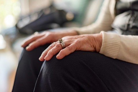 An elderly woman sits on a couch with her hands resting on her knees, wearing a ring on her left