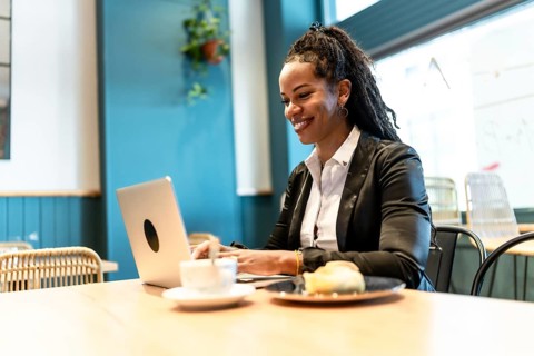 Confident latin businesswoman working with laptop in a cafeteria while eating sandwich and drinking