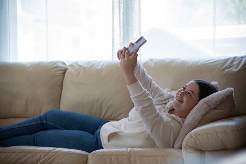 A girl lies comfortably on a sofa, smiling while using her smartphone in a naturally lit room