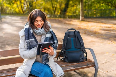 Caucasian smiling woman wearing warm clothes sitting on a park bench using digital tablet