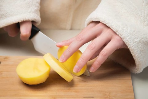 Young caucasian woman in a bathrobe stands in the kitchen and cuts fresh potatoes with a knife, on