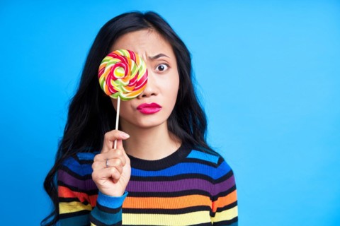 Confused thoughtful woman covers her eye with colorful lollipop looking to camera isolated on blue