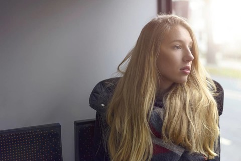 Young blond woman sitting in shadowed natural light looking out of a windows at the street outside