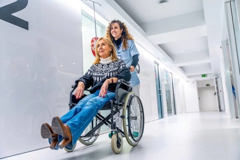 Nurse smiling while pushing a wheelchair with a injured woman along the corridor of the hospital