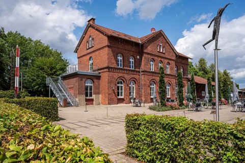 A red brick house with staircase and walkway, surrounded by green plants under a partly cloudy sky,