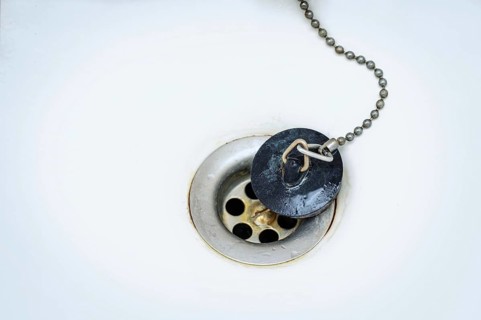 Old black rubber plug with metal chain on a rusty drain in a white wash basin or bathtub, copy
