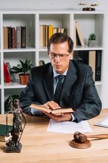 Mature male lawyer reading book with gavel justice statue wooden table