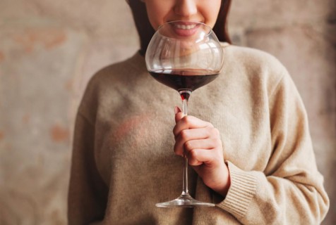 Crop happy female in sweater smelling red wine in large glass against gray background