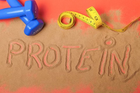 Word protein written on protein powder on red background, with dumbbells and measuring tape
