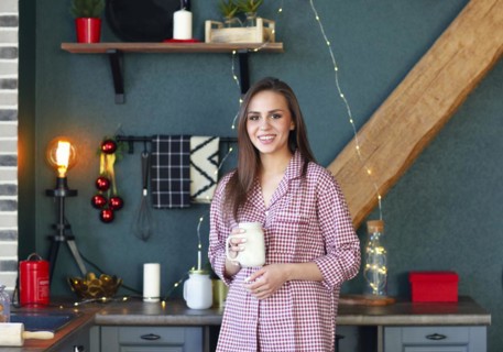 Happy young woman in plaid pajama holding jar smiling at camera in kitchen decorated for Xmas