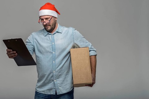 Cheerful attractive young man in a Santa Hat carrying or delivering a large brown cardboard box for