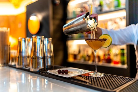 Hand of a waiter pouring cocktail into martini glass garnished with lemon in the counter of a