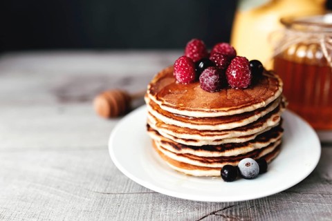 Juicy pancakes with berries and honey on a white plate, spoon, jar, wooden table, yellow vase with