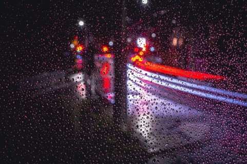 Rain falls on a pane of glass, blurred traces of light from vehicles and street lamps at night