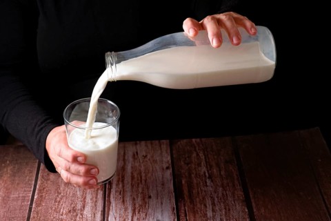 Hands of a woman pouring fresh milk into a glass, with a glass bottle
