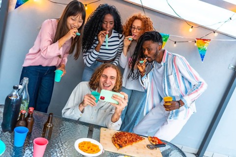 Happy caucasian man taking selfie surrounded by multi-ethnic friends blowing streamers during