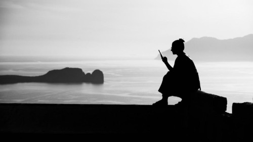 A woman sits on a stone wall and looks at her mobile phone, Venetian Castle, Plaka, Milos,