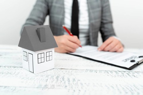 Businesswoman sitting and writing in notebook. Paper house on desk
