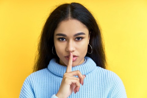 Pretty black woman keeps finger on lips, making hush gesture on yellow background. Shh, silence