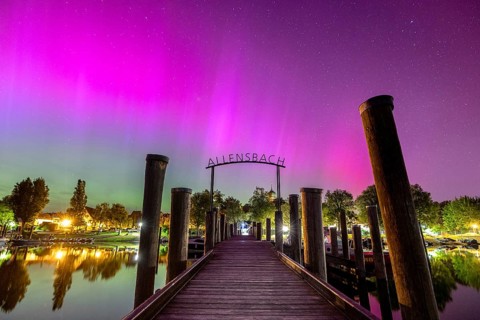 A wooden walkway under a glowing sky with colourful lights and trees with auroras, harbour,
