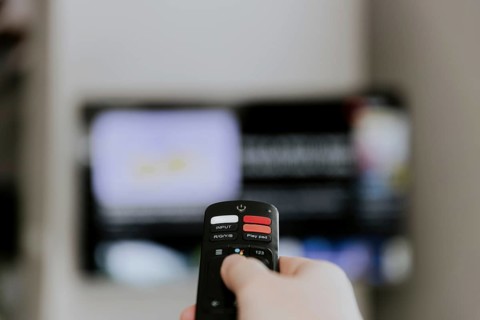 Hand holding remote control pointing to TV. Person watching smart television indoors. Blurred
