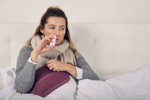Sick woman wrapped in a warm scarf and clutching a hot water bottle sitting using nasal spray with