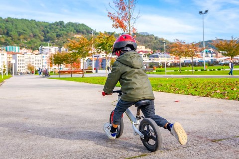 A young boy is riding a bicycle down a sidewalk. He is wearing a helmet and a green jacket