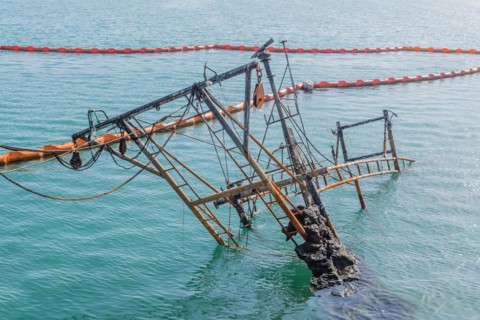 Rusty remnants of a sunken ship with booms and an orange barrier on calm ocean water, in Sinjin-do,