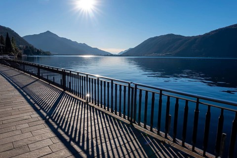Shadow of a Railing on the Street on the Waterfront to Lake Ceresio with Mountain and Sunbeam in