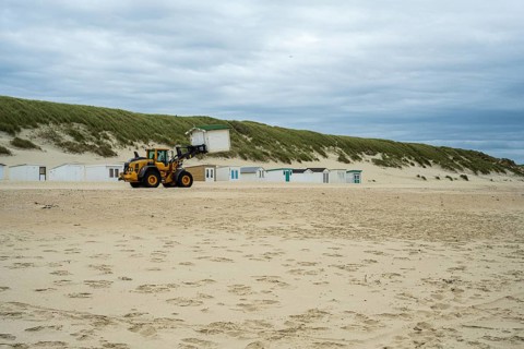 A tractor transports a beach cabin across an empty beach with dunes in the background, end of
