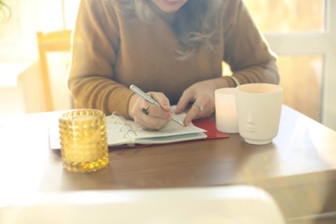 Middle-aged woman writer or poet writing in notebook while sitting at table in sunny room at home,