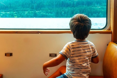 A young boy is sitting on a train window looking out at the water. The boy is wearing a striped