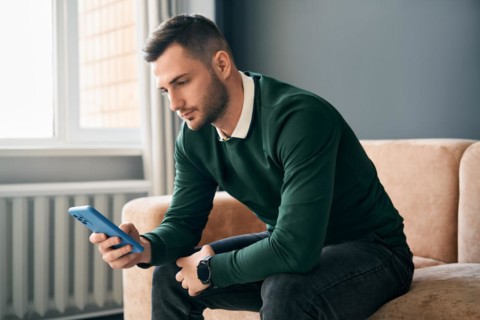 Relaxed man sitting on couch using hsi mobile smart phone at home. Social media concept