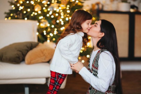 Beautiful loving mother gently kissing cute little daughter against festively decorated Christmas