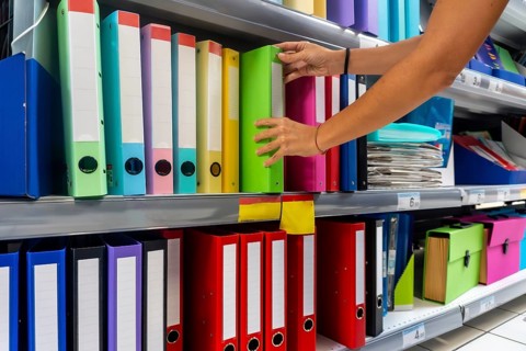 A person is reaching for a green binder on a shelf. The shelf is full of colorful binders and