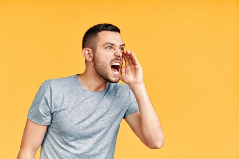 Young angry man shouting and screaming loud to side with hand on mouth and copy space for text over