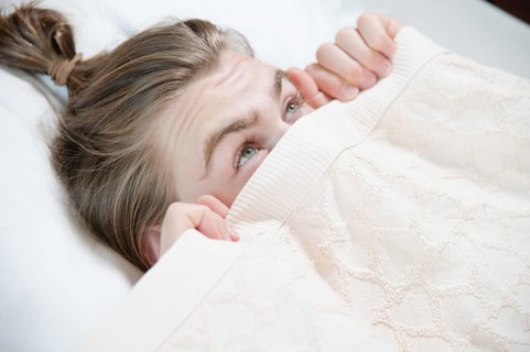 A young attractive man peeks out from under the blankets hiding behind them with fear when he wakes