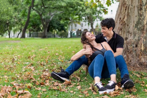 Couple sitting in a park leaning on a tree. Happy lovers laughing