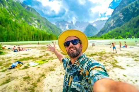 Happy man in a hawaiian shirt and straw hat taking a selfie at lago di landro, with tourists and