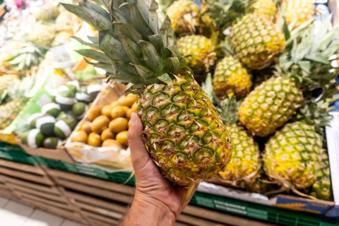 A person is holding a pineapple in a grocery store. The pineapple is surrounded by other fruits and