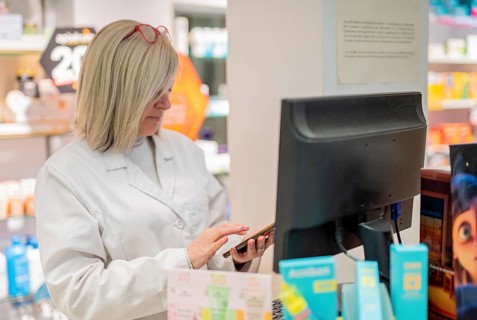 Efficient blonde pharmacist updates stock with smartphone and computer in a tidy pharmacy.