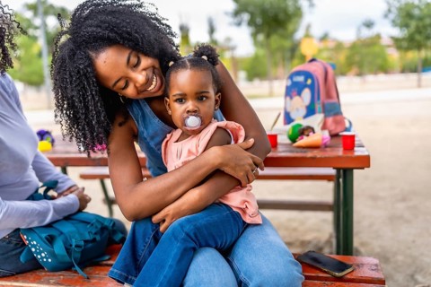Happy african mother sitting on picnic table embracing a baby girl with pacifier in the park