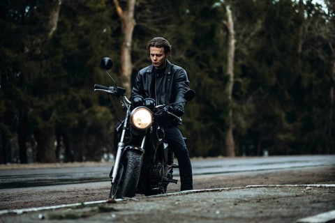 Stylish male motorcyclist in a leather jacket with a classic motorcycle on a forest asphalt road