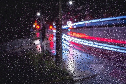 Rainy window pane with blurred light traces of passing vehicles in the dark, urban atmosphere,