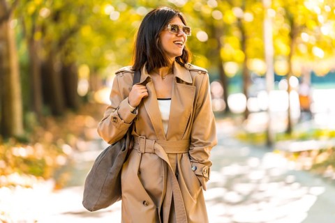 A woman wearing a tan trench coat and sunglasses walks down a sidewalk. She is smiling and carrying