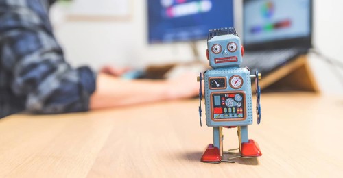 Toy robot on a desktop, man working on the computer in the background. Symbol for artificial
